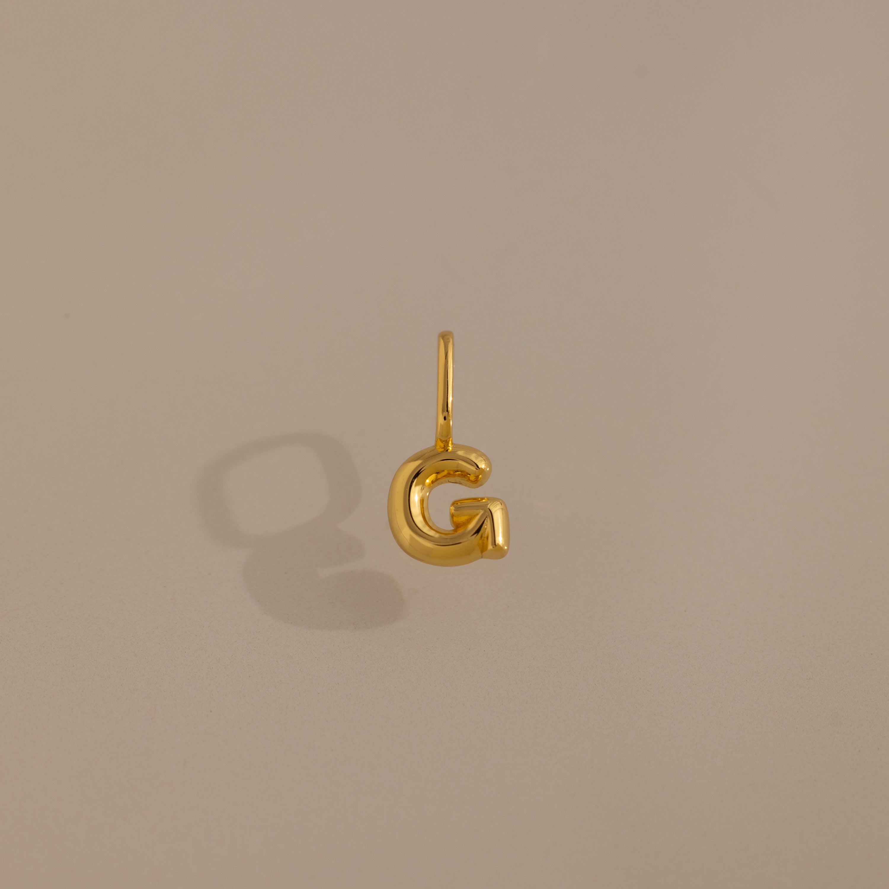 The Bubble Letter Charm in 18K Gold, shaped as the letter G, offers a stylish personalized jewelry option and is showcased on a simple beige background.