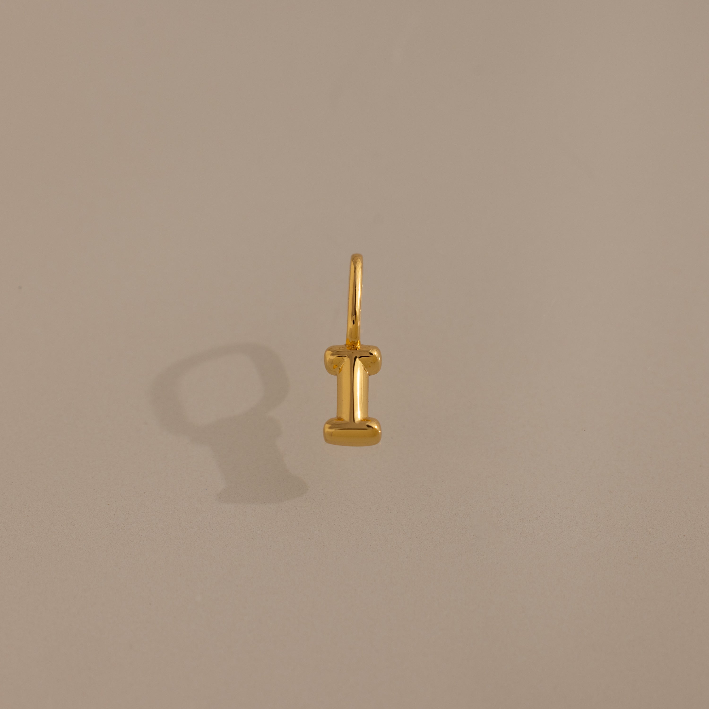 The Bubble Letter Charm in 18K Gold, shaped like the letter I, rests on a plain beige background—an elegant piece of personalized jewelry.
