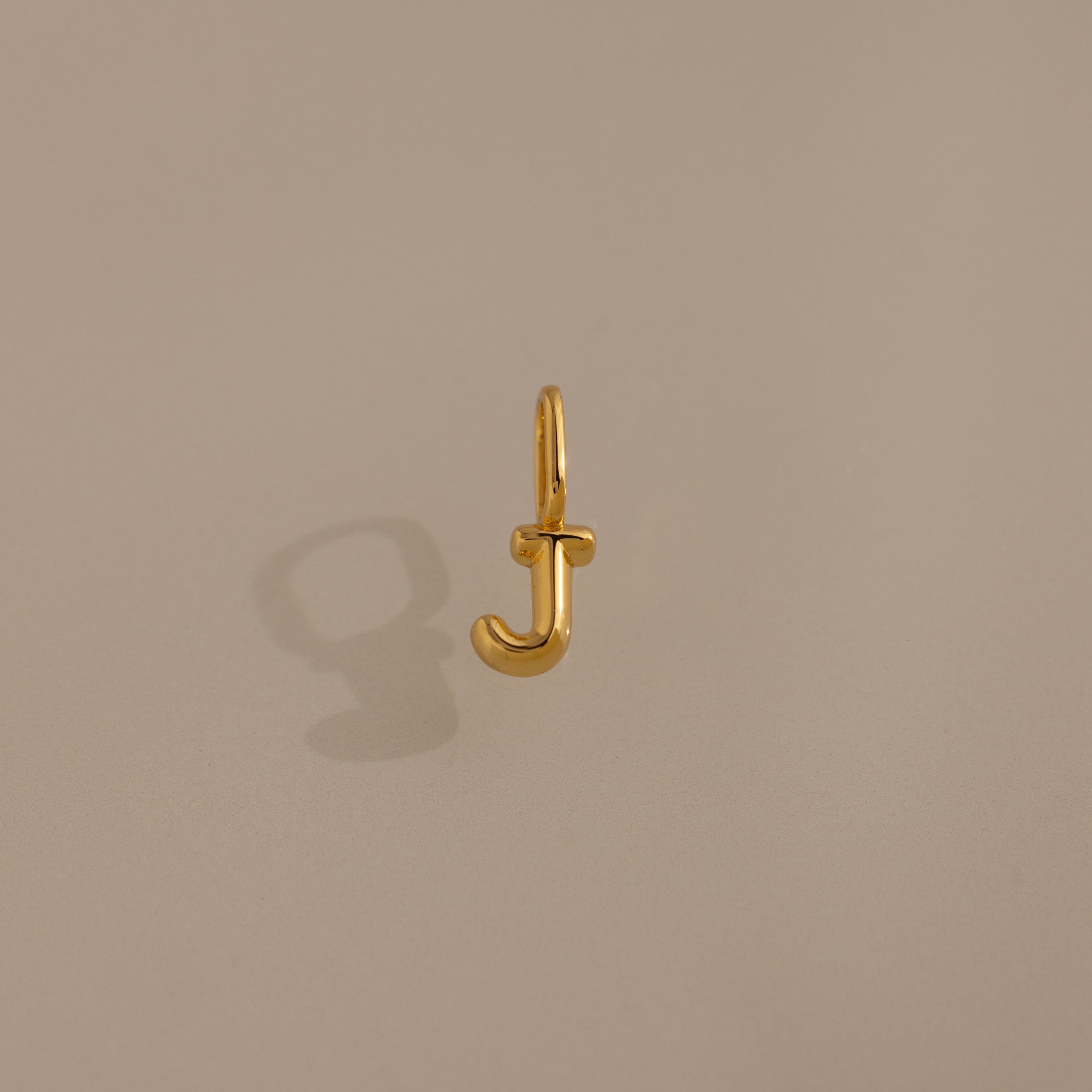 The Bubble Letter Charm in 18K Gold, shaped like the letter J and photographed on a plain beige background, makes a perfect personalized jewelry piece or a charming addition to your bracelet.