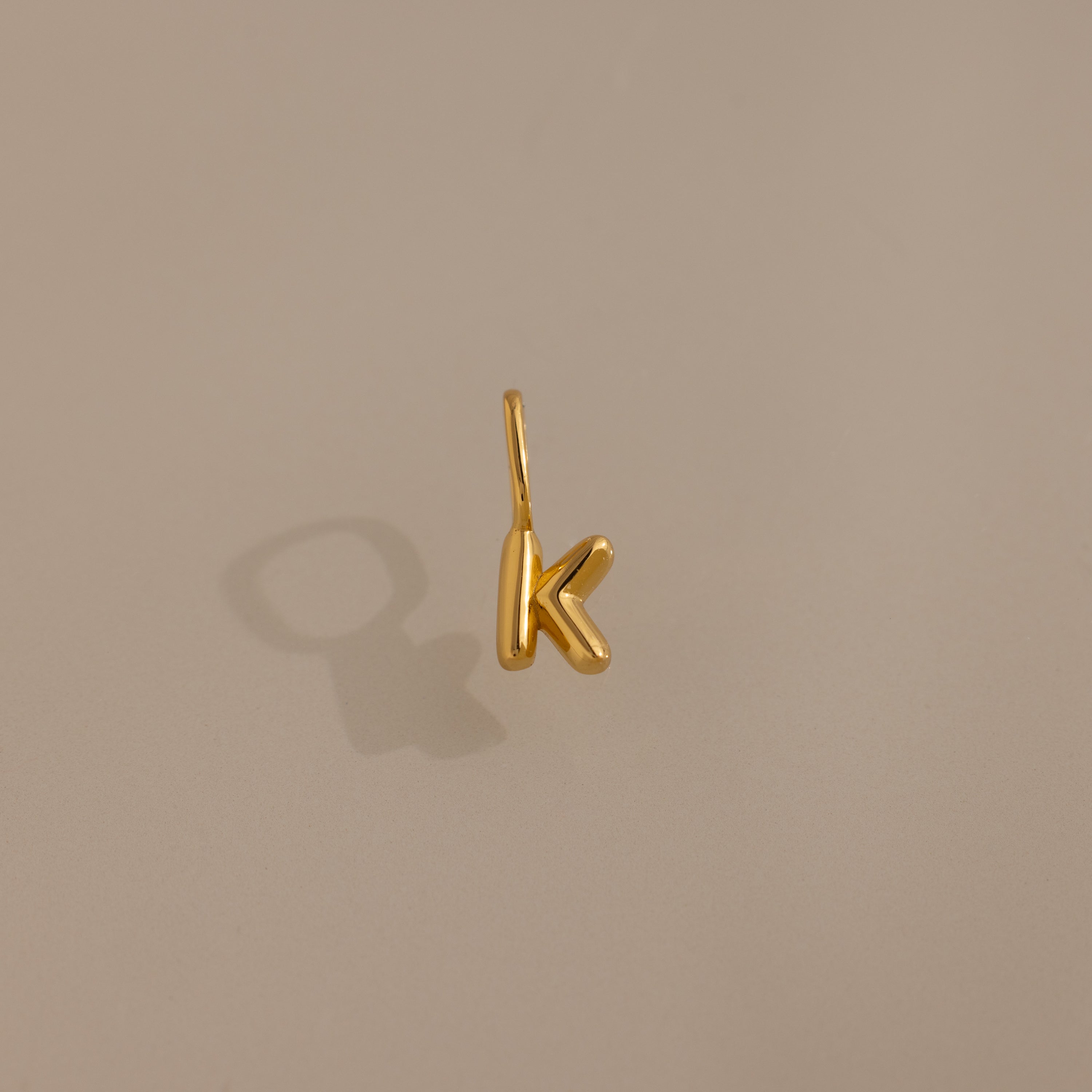 The Bubble Letter Charm in 18K Gold, shaped as a K, makes a stylish personalized pendant or bracelet charm and is showcased on a neutral beige background with a soft shadow.