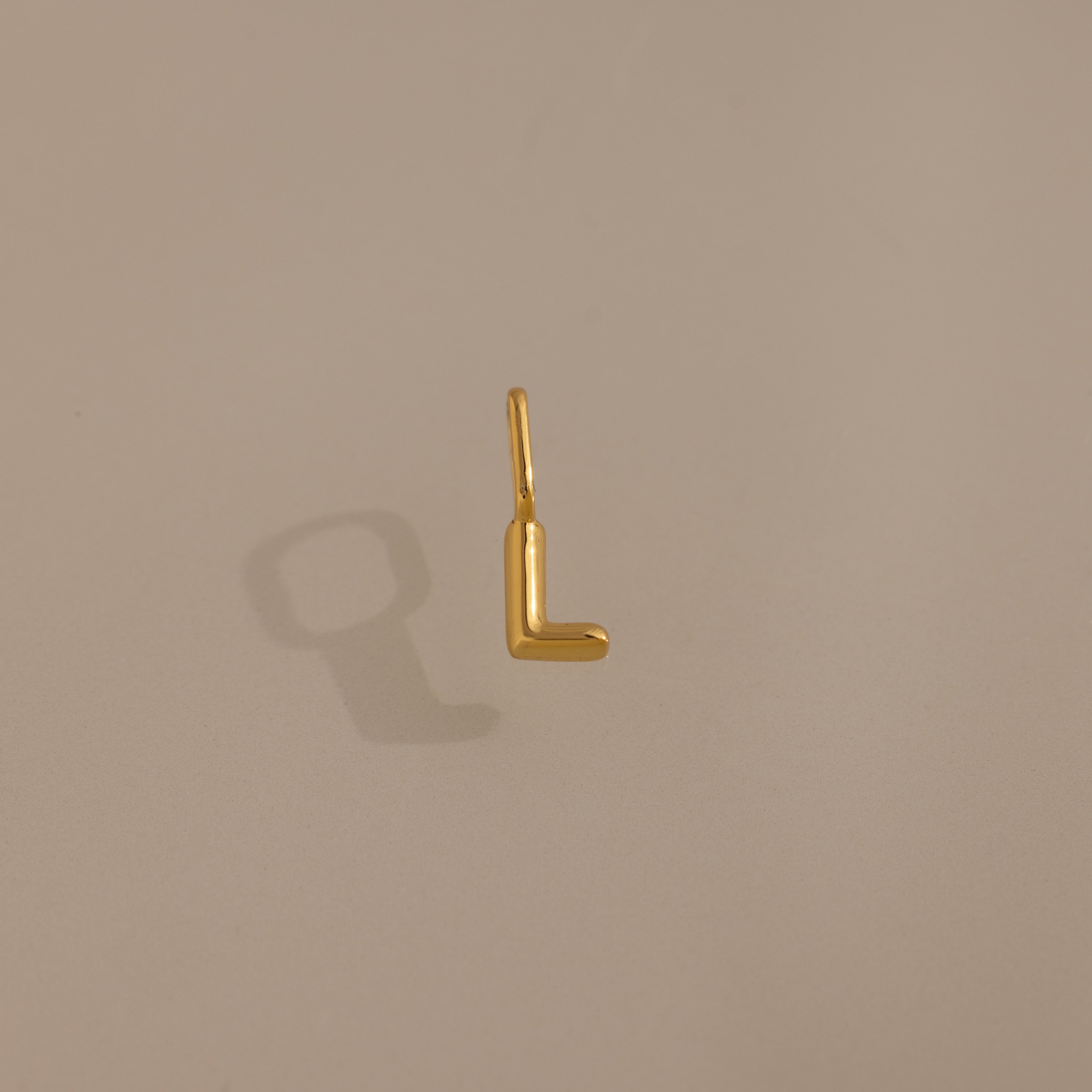The Bubble Letter Charm in 18K Gold, shaped like an 'L', casts a shadow on a neutral background—a stylish, personalized jewelry piece ideal as a charm for a bracelet or necklace.