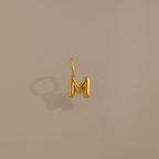 Bubble Letter Charm in 18K Gold—a gold bubble letter M charm for bracelets, displayed on a beige background with a soft left-side shadow. Ideal for adding personalized style to your jewelry collection.