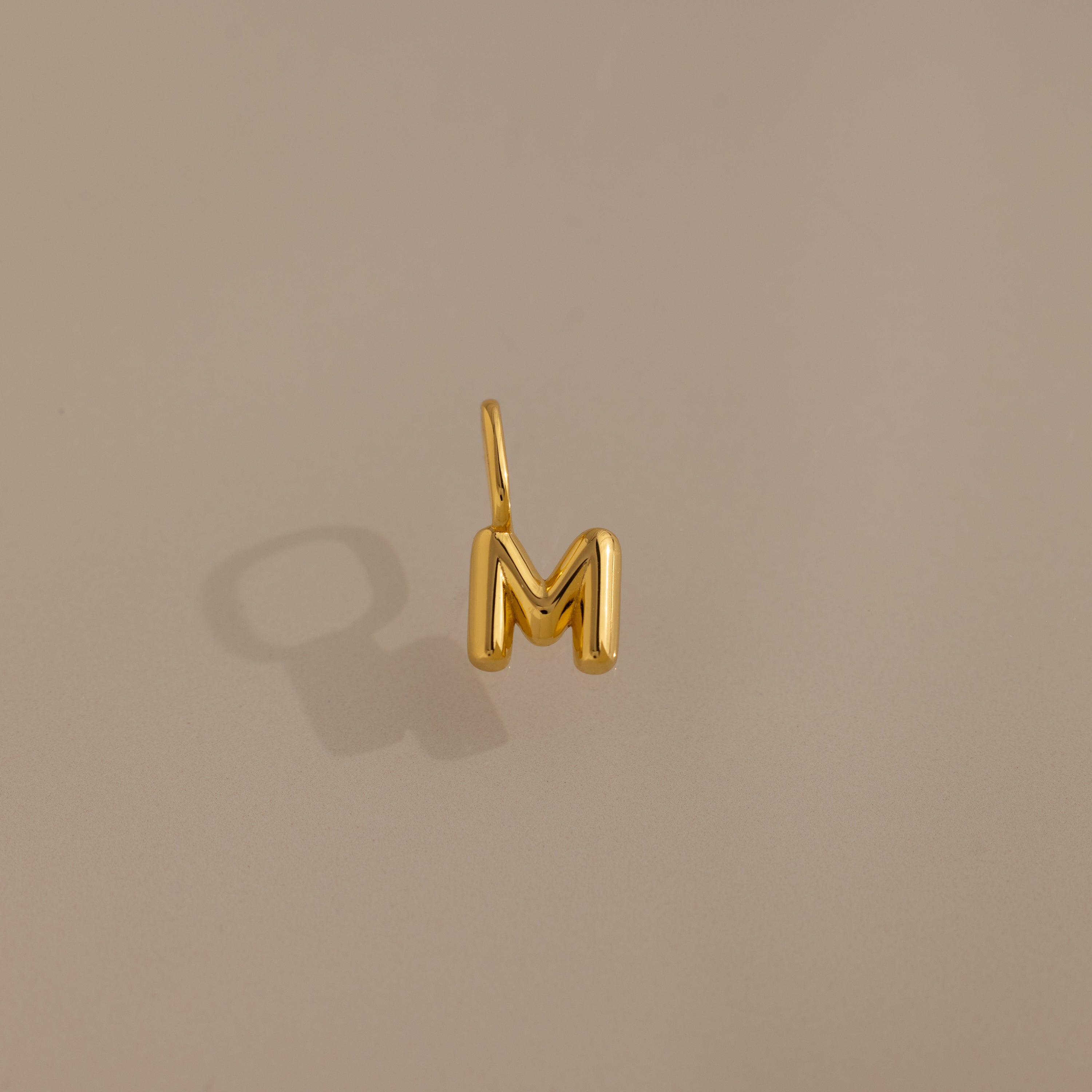 Bubble Letter Charm in 18K Gold—a gold bubble letter M charm for bracelets, displayed on a beige background with a soft left-side shadow. Ideal for adding personalized style to your jewelry collection.