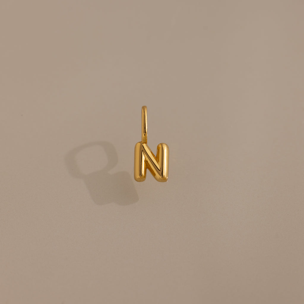 Bubble Letter Charm in 18K Gold shaped as the letter N, displayed on a plain beige background with a shadow. A stylish personalized jewelry piece to enhance your collection or add as a charm for your bracelet.