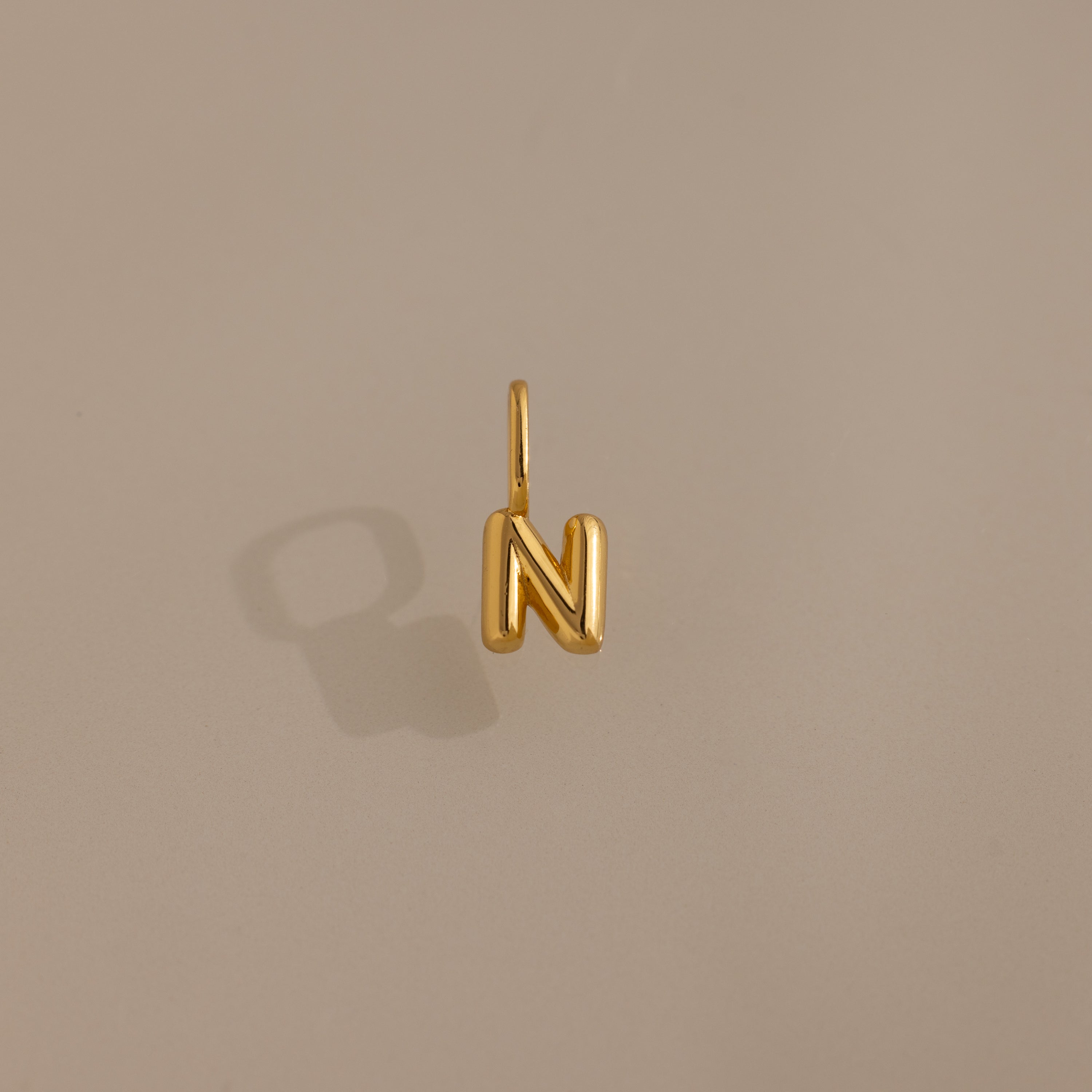 Bubble Letter Charm in 18K Gold shaped as the letter N, displayed on a plain beige background with a shadow. A stylish personalized jewelry piece to enhance your collection or add as a charm for your bracelet.
