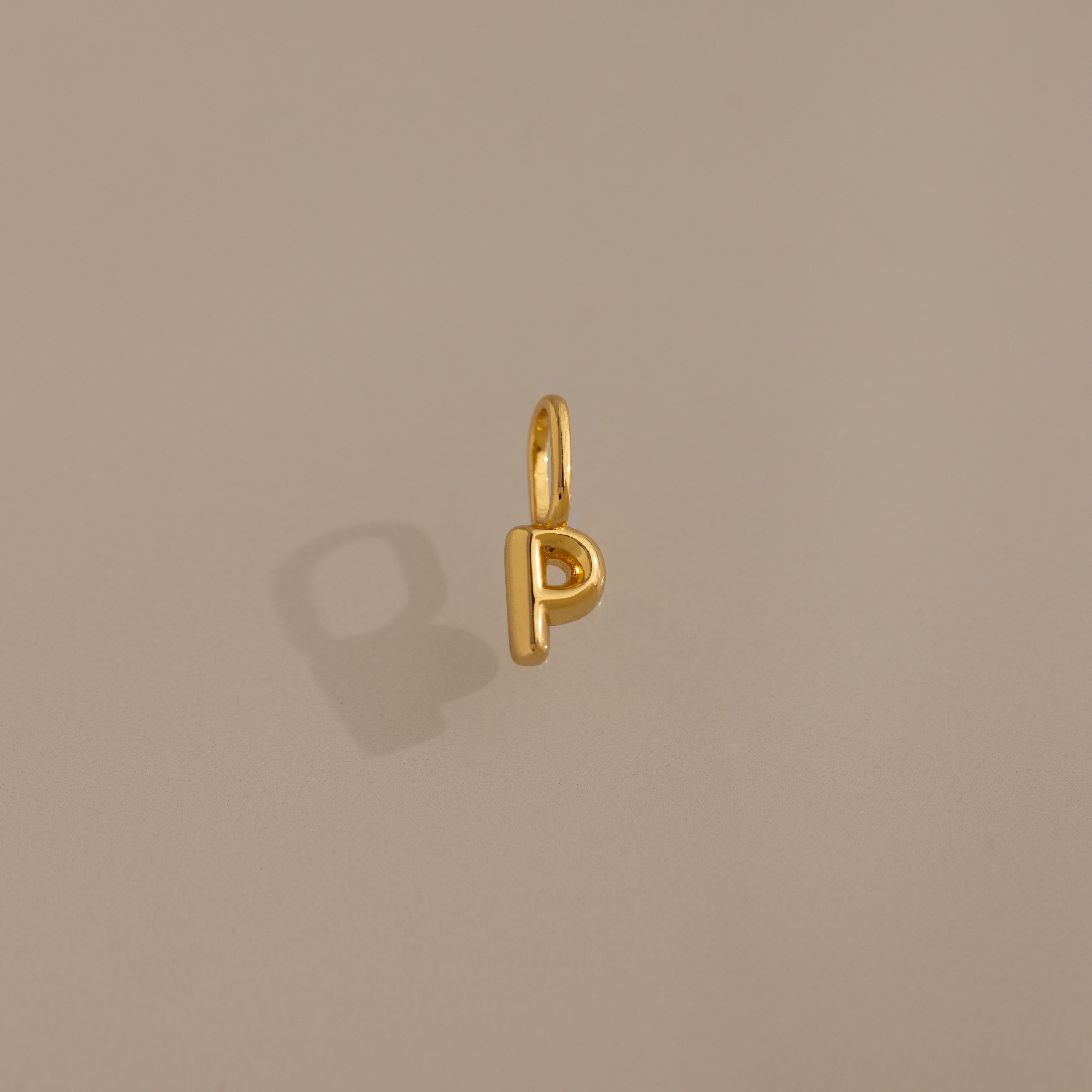 The Bubble Letter Charm in 18K Gold shaped like the letter P rests on a plain beige background, making it an ideal personalized jewelry piece or trendy charm for bracelet lovers.