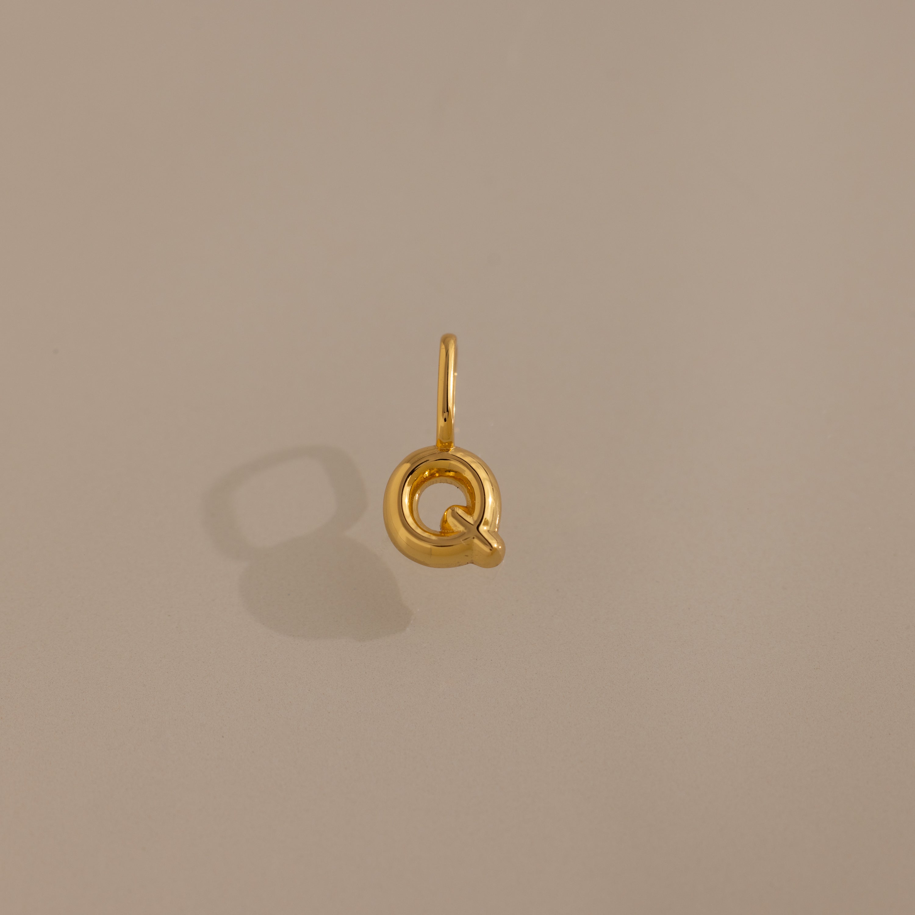 The Bubble Letter Charm in 18K Gold is a circular gold pendant with a small loop, shown on a beige background with a soft shadow—ideal for personalized jewelry or bracelet lovers.