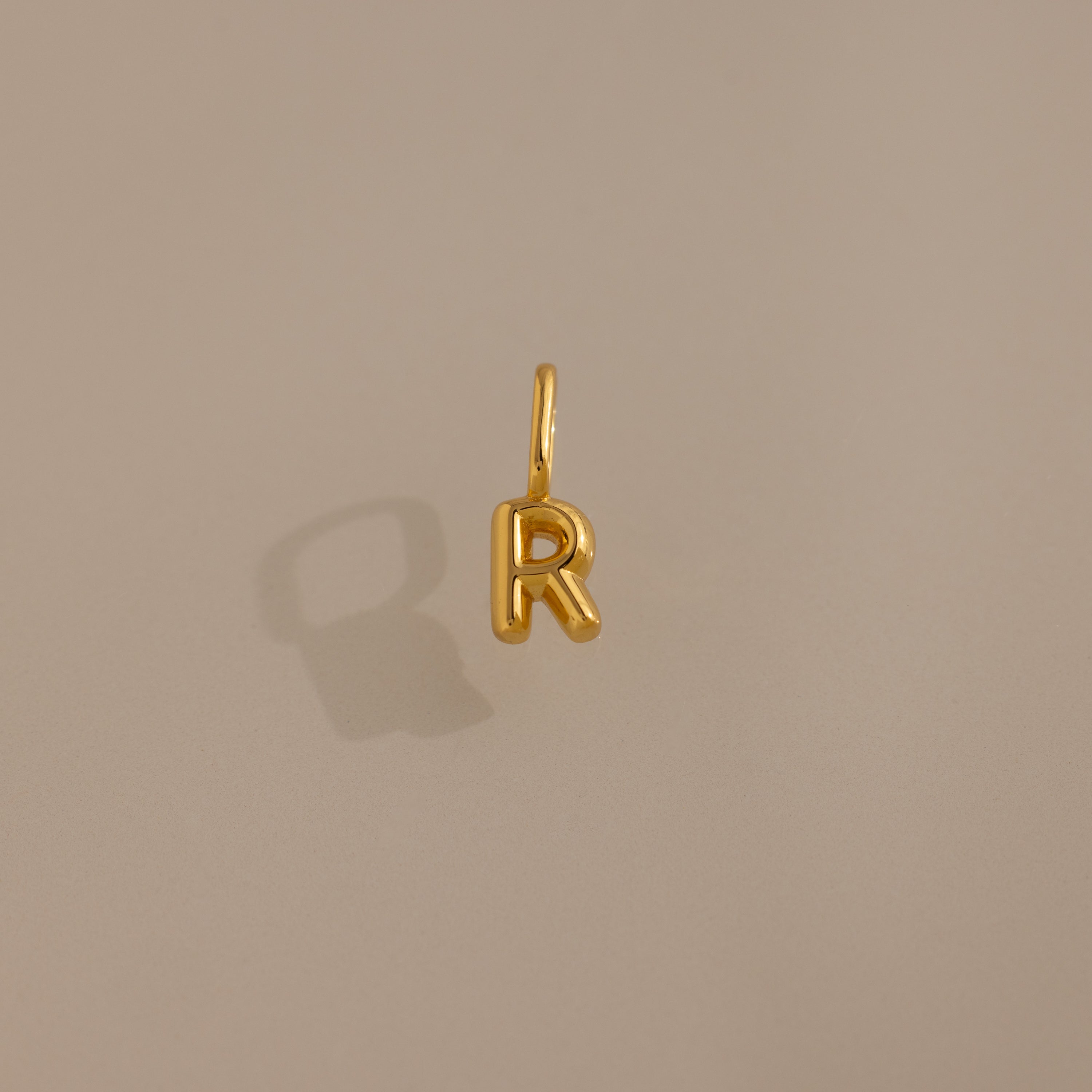 The Bubble Letter Charm in 18K Gold is a playful R-shaped pendant on a plain beige background with a subtle shadow—ideal as personalized jewelry or a unique charm for bracelet lovers.