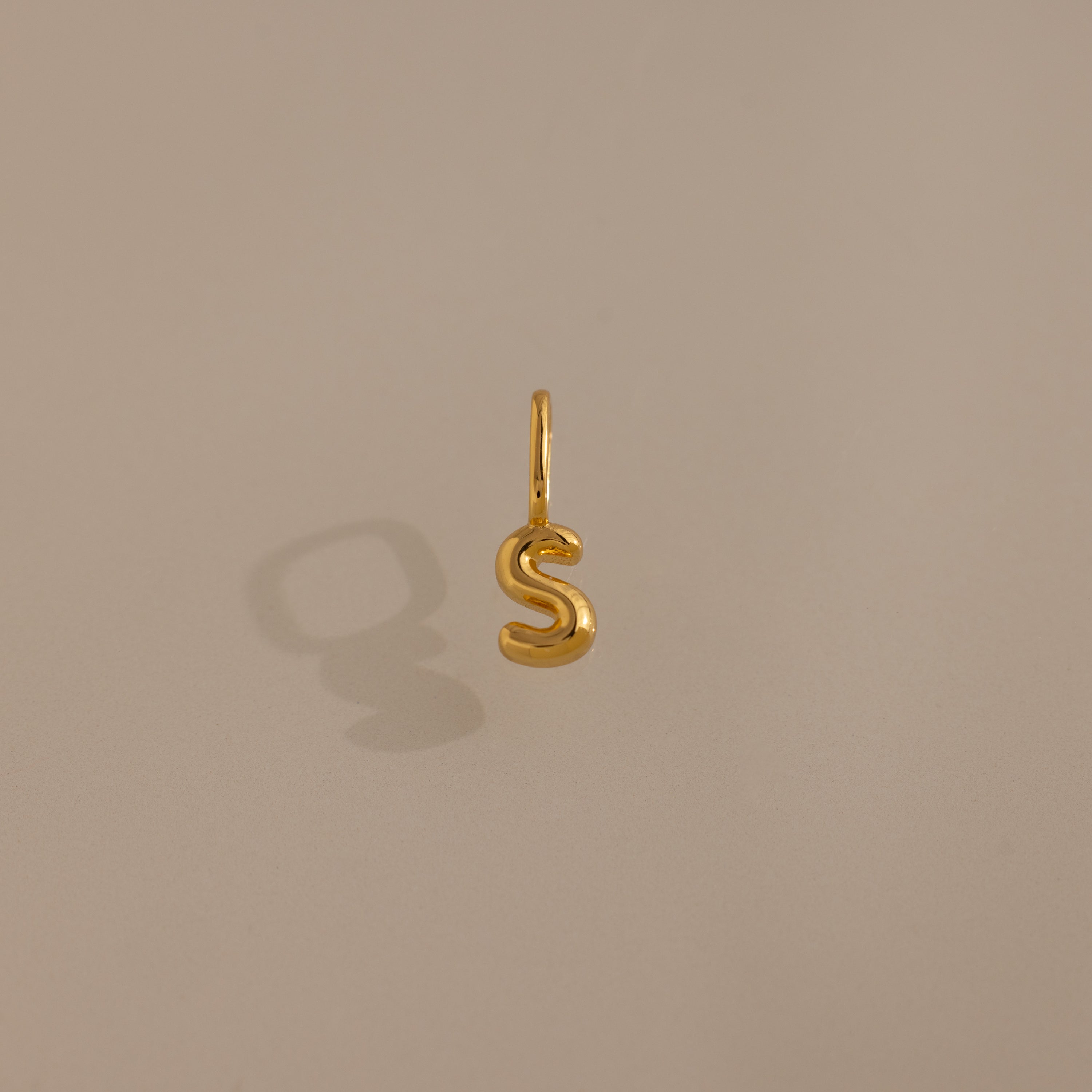 The Bubble Letter Charm in 18K Gold casts a shadow on a plain beige surface, making it a standout accessory—ideal as personalized jewelry or a unique addition to bracelet collections.