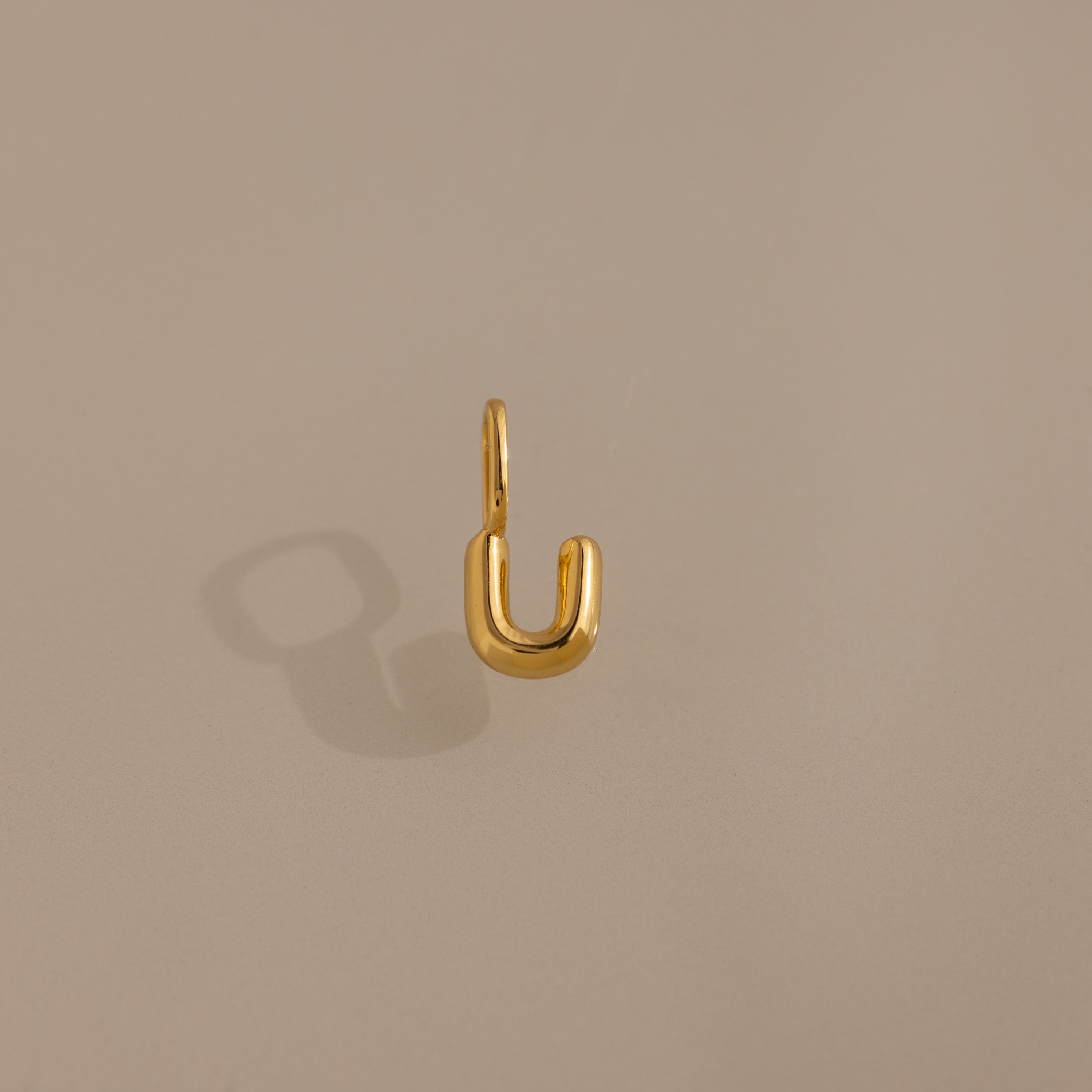 The Bubble Letter Charm in 18K Gold forms a striking U-shape on a beige surface, making it an ideal personalized jewelry piece or charm for bracelet lovers.