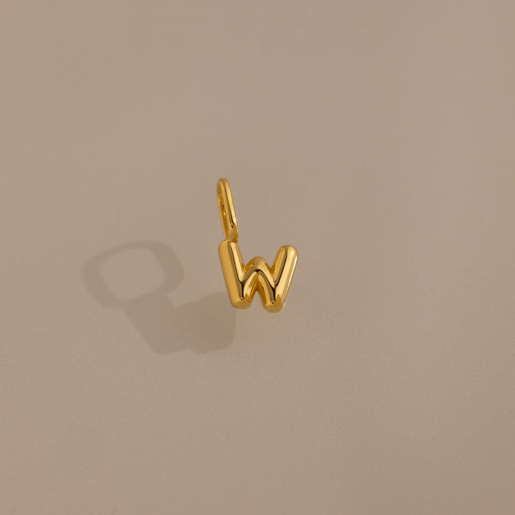 The Bubble Letter Charm in 18K Gold, shaped like the letter W, sits on a soft beige background with a subtle shadow—ideal as personalized jewelry or a stylish charm for bracelet enthusiasts.