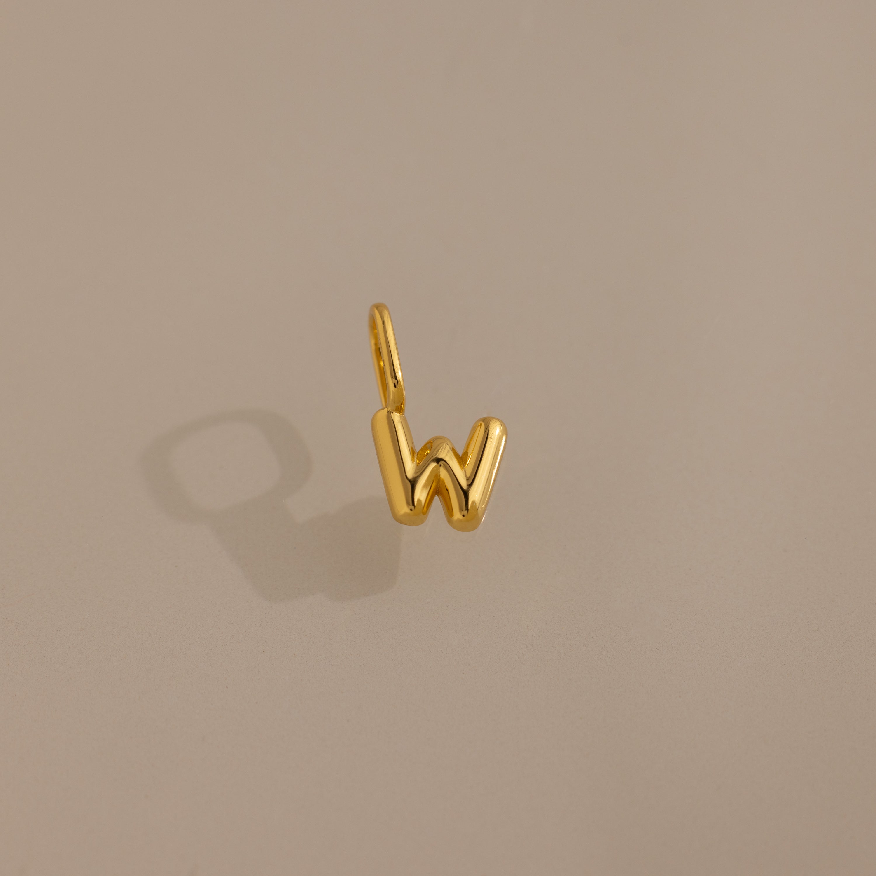 The Bubble Letter Charm in 18K Gold, shaped like the letter W, sits on a soft beige background with a subtle shadow—ideal as personalized jewelry or a stylish charm for bracelet enthusiasts.