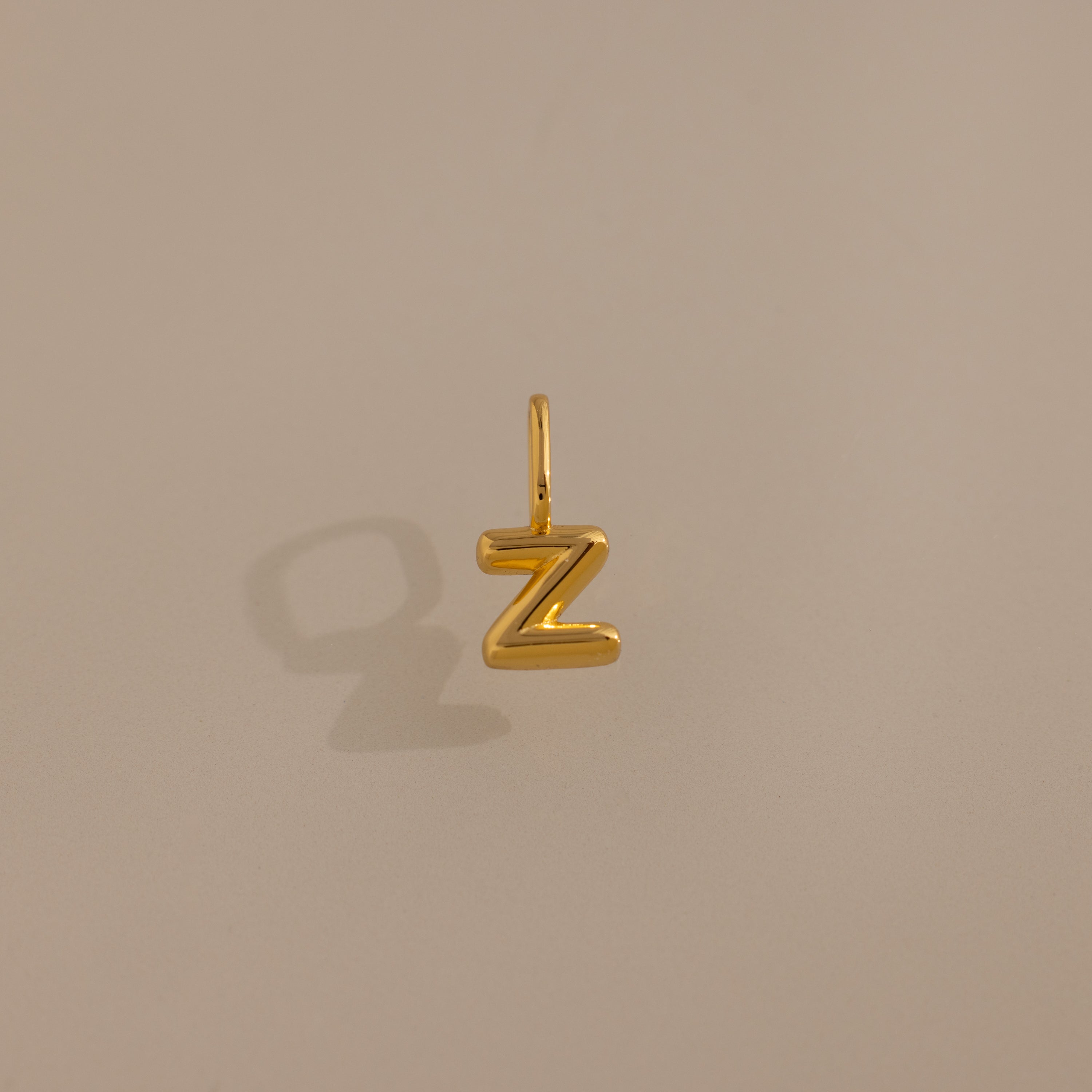 The Bubble Letter Charm in 18K Gold, shaped like a Z and casting a shadow on a beige surface, makes for a standout piece of personalized jewelry.