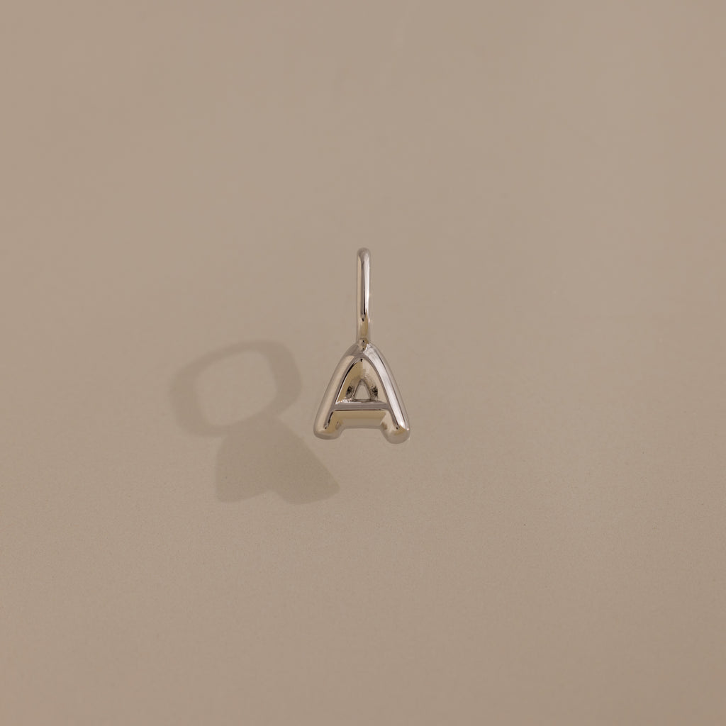 The Bubble Letter Charm in Sterling Silver, shaped like the letter A, rests on a beige background with a soft shadow—an ideal choice for personalized jewelry fans.