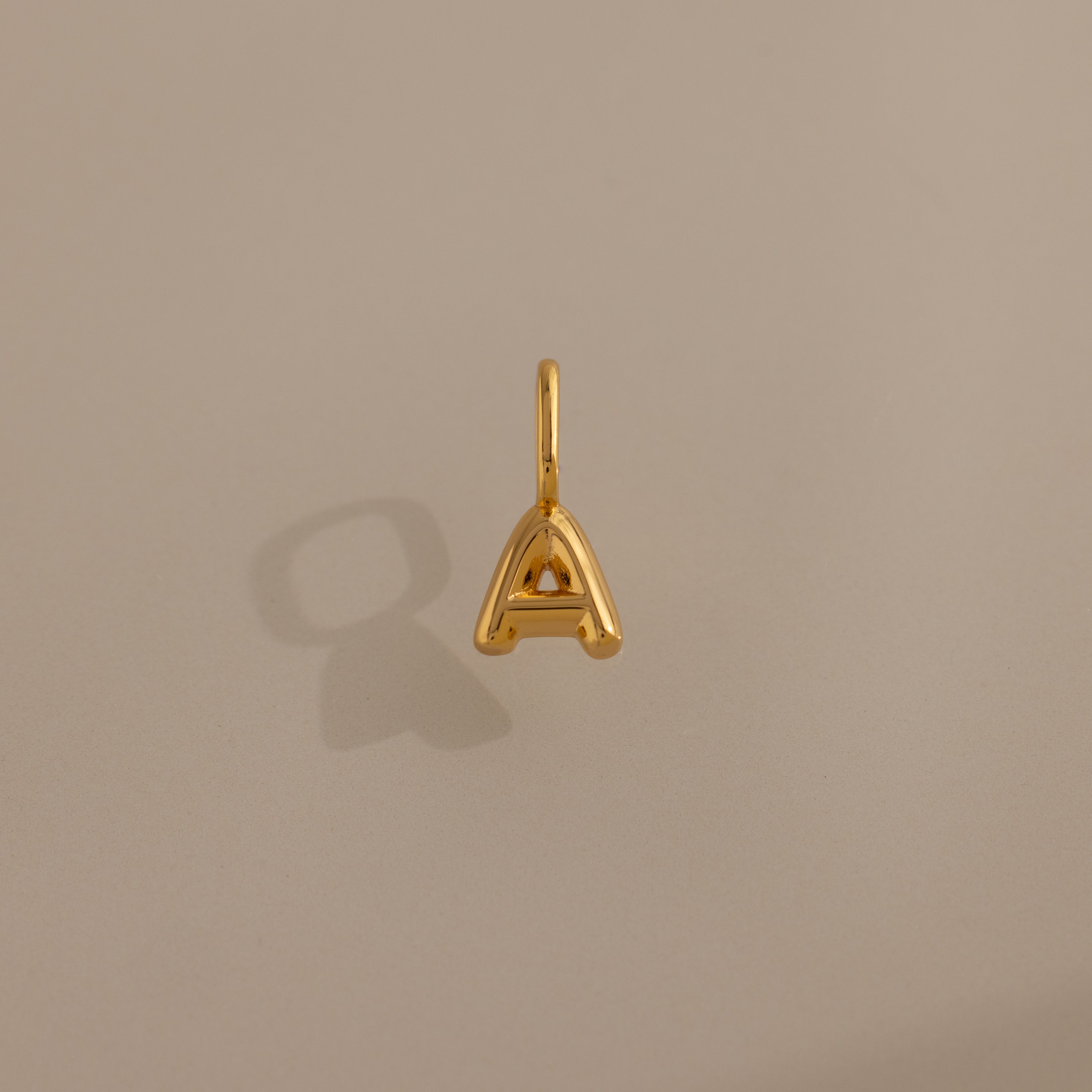 Bubble Letter Charm in 18K Gold shaped as an "A" shown on a plain beige background, casting a subtle shadow to the left.