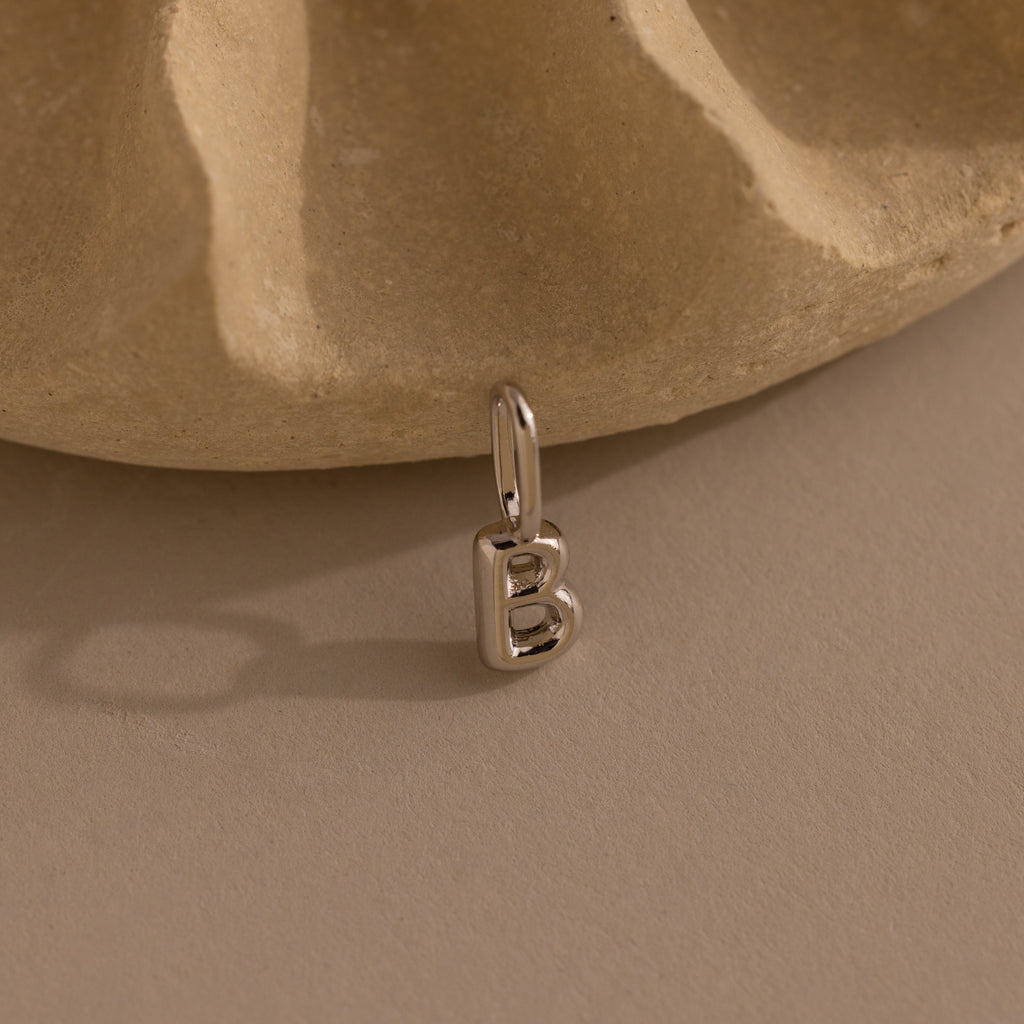 Silver bubble letter “B” charm resting upright against a light stone background. The charm has a rounded, dimensional shape and a loop at the top for attaching.