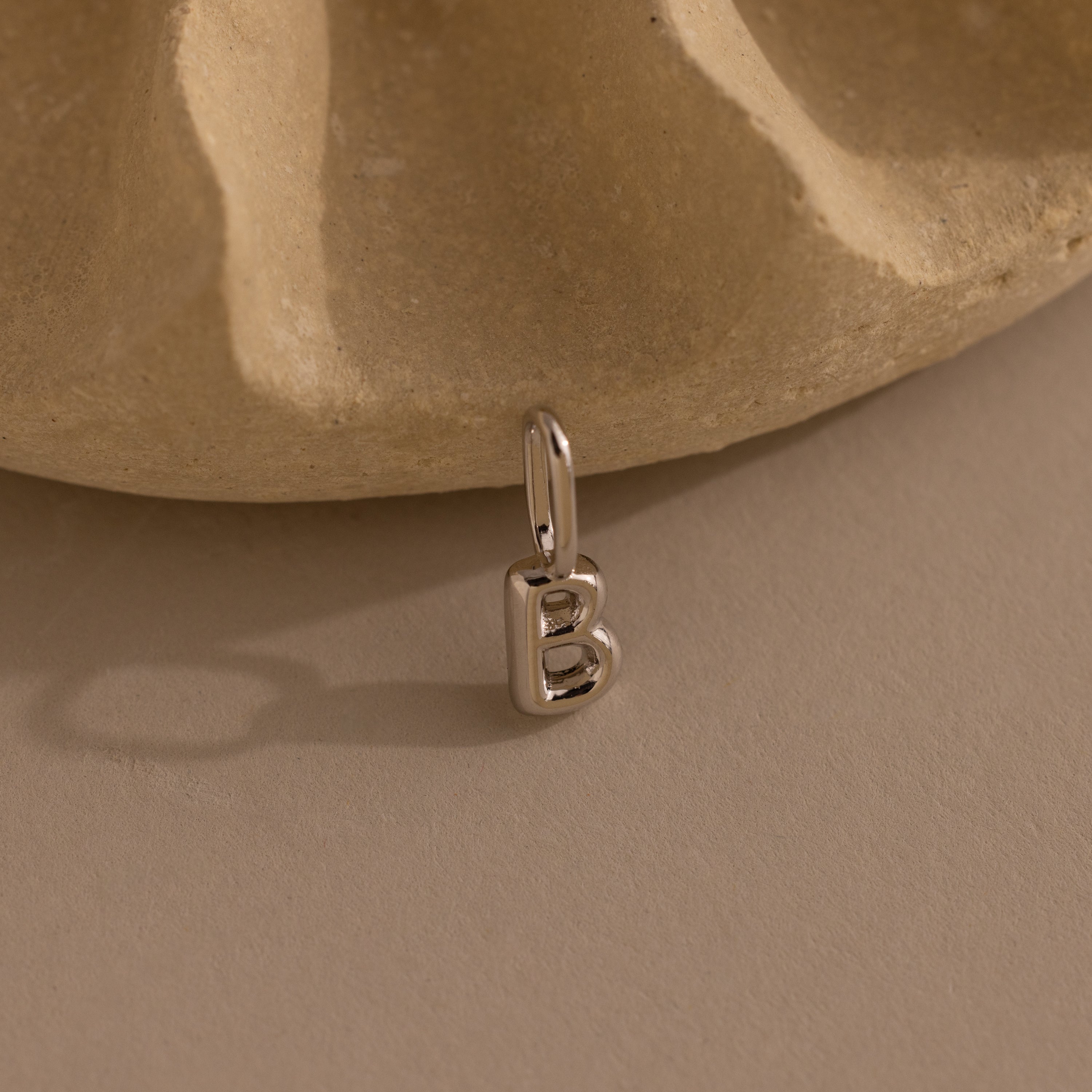 Silver bubble letter “B” charm resting upright against a light stone background. The charm has a rounded, dimensional shape and a loop at the top for attaching.