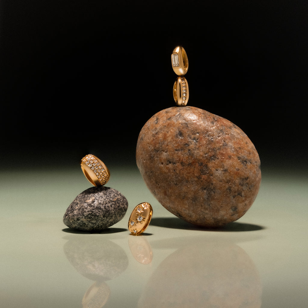 The Bold Pave Dome Charm in 18K Gold is elegantly showcased on and around smooth stones, creating a striking visual against a dark gradient background—an ideal presentation for fine jewelry.