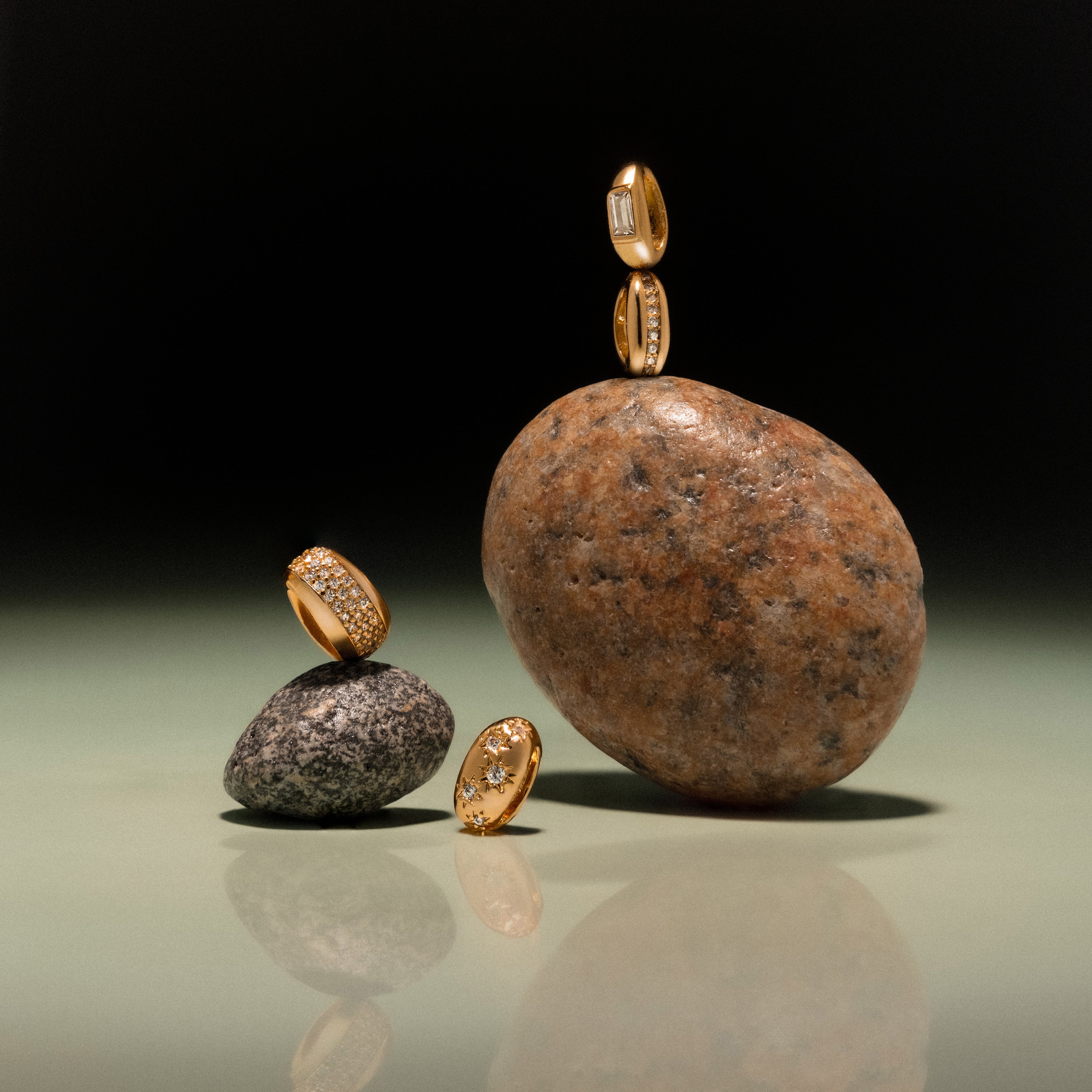 The Bold Pave Dome Charm in 18K Gold is elegantly showcased on and around smooth stones, creating a striking visual against a dark gradient background—an ideal presentation for fine jewelry.