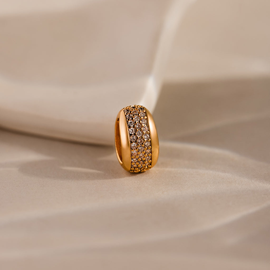 The Bold Pave Dome Charm in 18K Gold features a wide, sparkling band and rests on a soft beige surface—an ideal statement piece for any elegant occasion.