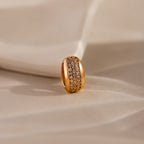 The Bold Pave Dome Charm in 18K Gold features a wide, sparkling band and rests on a soft beige surface—an ideal statement piece for any elegant occasion.
