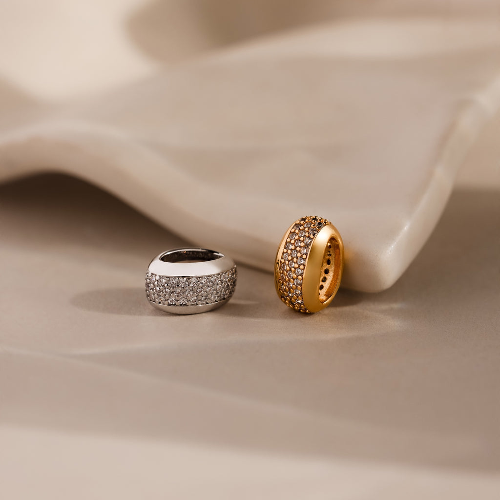The Bold Pave Dome Charm, featuring both silver and gold tones with shimmering embedded stones, is elegantly displayed on a soft beige surface—an ideal choice for jewelry lovers seeking timeless ring designs.
