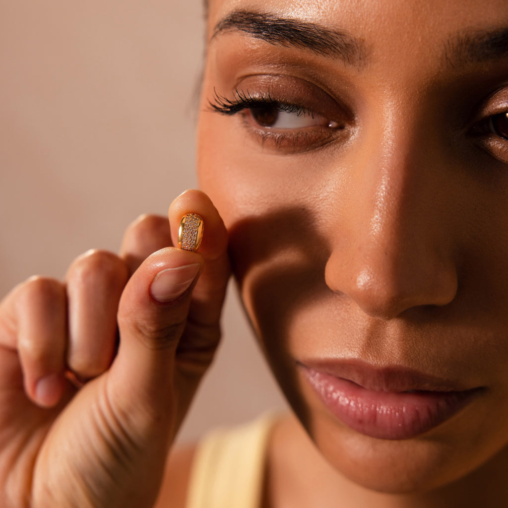 A woman holds the Bold Pave Dome Charm in 18K Gold between her fingers near her face, examining it closely.