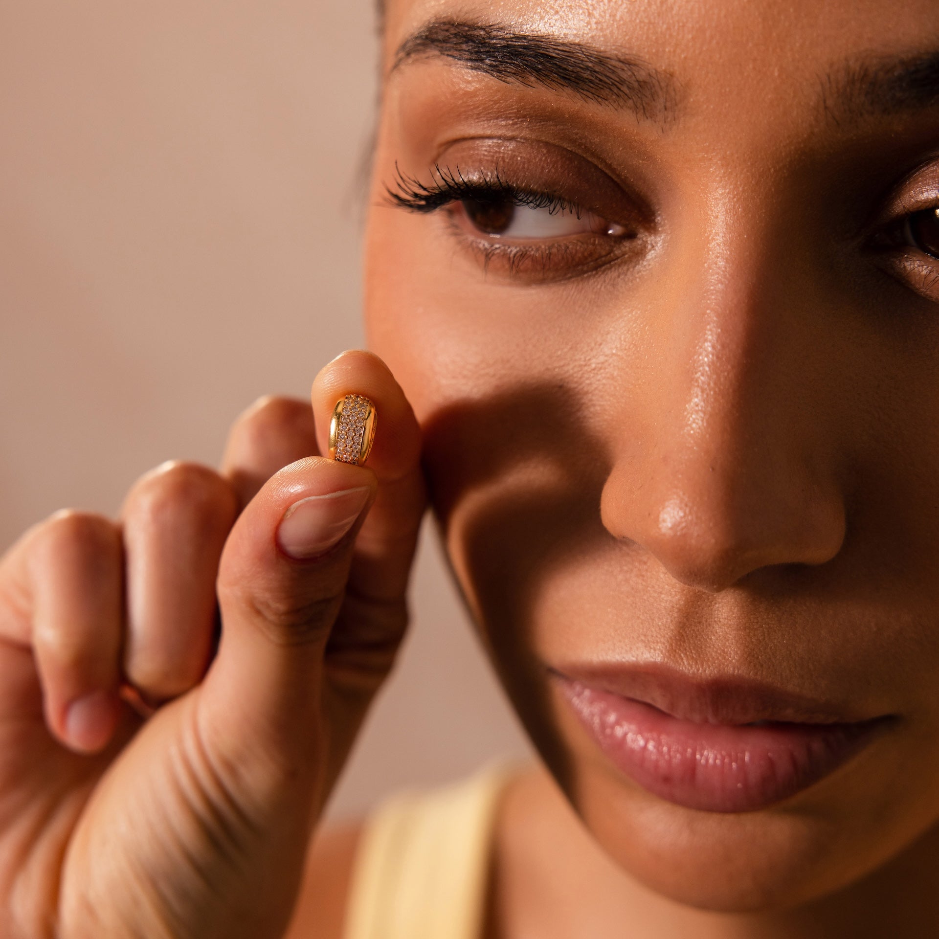 A woman holds the Bold Pave Dome Charm in 18K Gold between her fingers near her face, examining it closely.
