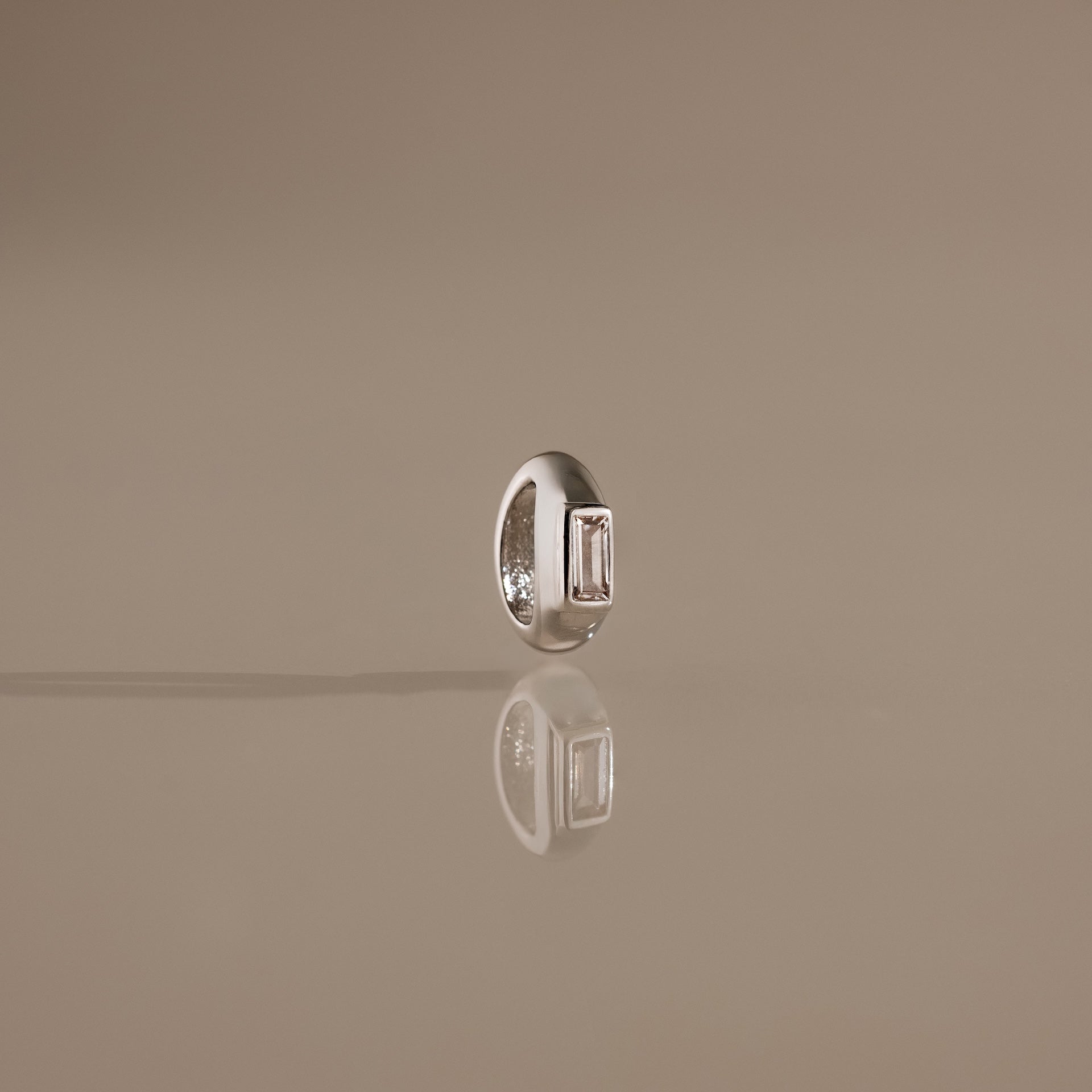 The Baguette Diamond Charm in Silver stands upright on a reflective beige surface, catching the light with its elegant rectangular gemstone and modern design.
