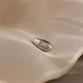The Baguette Diamond Charm in Silver, featuring a rectangular clear stone, rests elegantly on a smooth beige surface, making it a timeless jewelry piece.