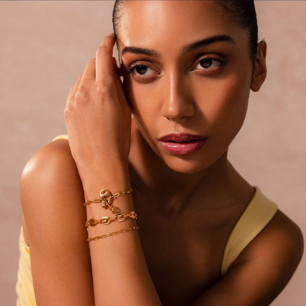 A woman in a yellow top wears Connector Clasp gold chain bracelets, touching her face and looking to the side—offering effortless elegance for anyone seeking standout jewelry.