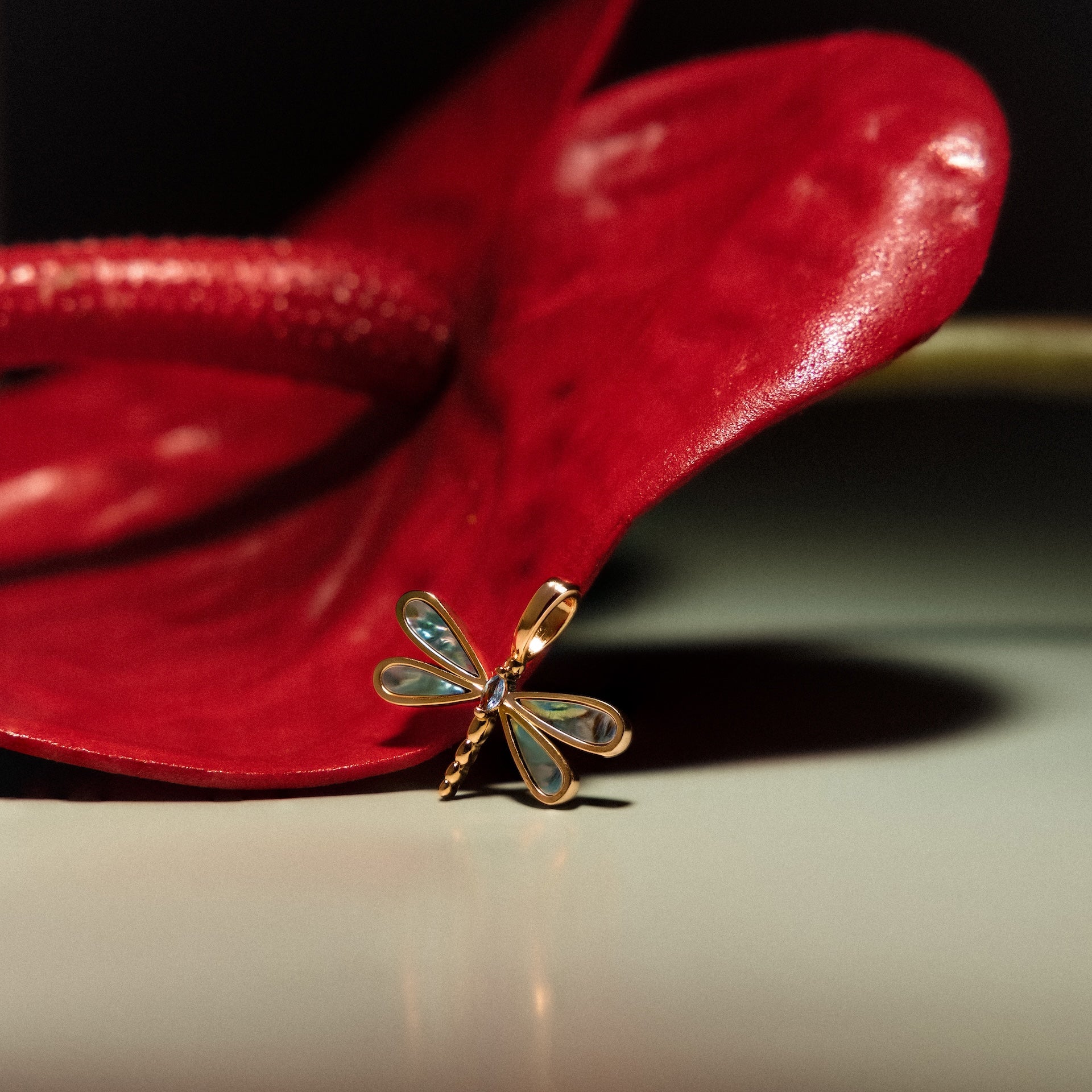 The Abalone Dragonfly Charm in 18K Gold, with iridescent details, rests on a vibrant red anthurium flower.