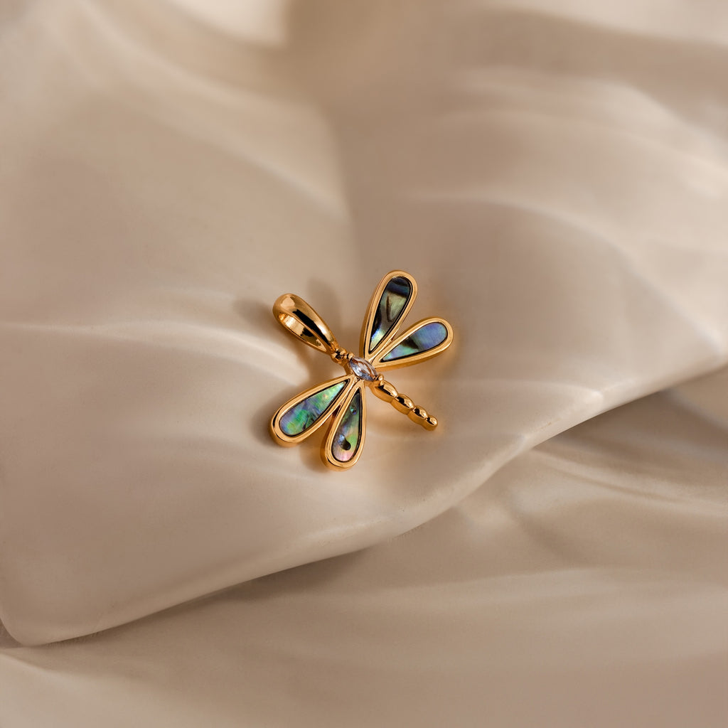 The Abalone Dragonfly Charm in 18K Gold, featuring iridescent wings, rests on soft cream fabric—a stunning nature-inspired accessory.