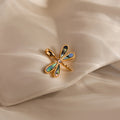 The Abalone Dragonfly Charm in 18K Gold, featuring iridescent wings, rests on soft cream fabric—a stunning nature-inspired accessory.