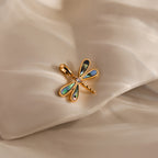 The Abalone Dragonfly Charm in 18K Gold, featuring iridescent wings, rests on soft cream fabric—a stunning nature-inspired accessory.