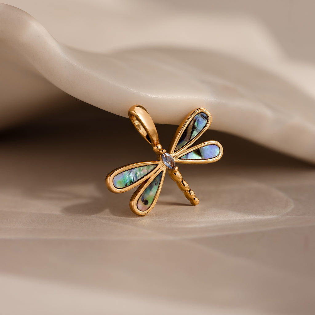 Abalone Dragonfly Charm in 18K Gold featuring blue and green iridescent wing details, displayed on a beige wavy surface—a nature-inspired accessory.