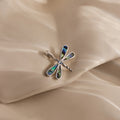 The Abalone Dragonfly Charm in Silver features iridescent wing details and is displayed on a silky, beige fabric background—a nature-inspired piece perfect for jewelry lovers.