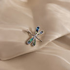 The Abalone Dragonfly Charm in Silver features iridescent wing details and is displayed on a silky, beige fabric background—a nature-inspired piece perfect for jewelry lovers.