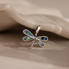 The Abalone Dragonfly Charm in Silver features iridescent wings and rests gracefully on a neutral, wavy surface.
