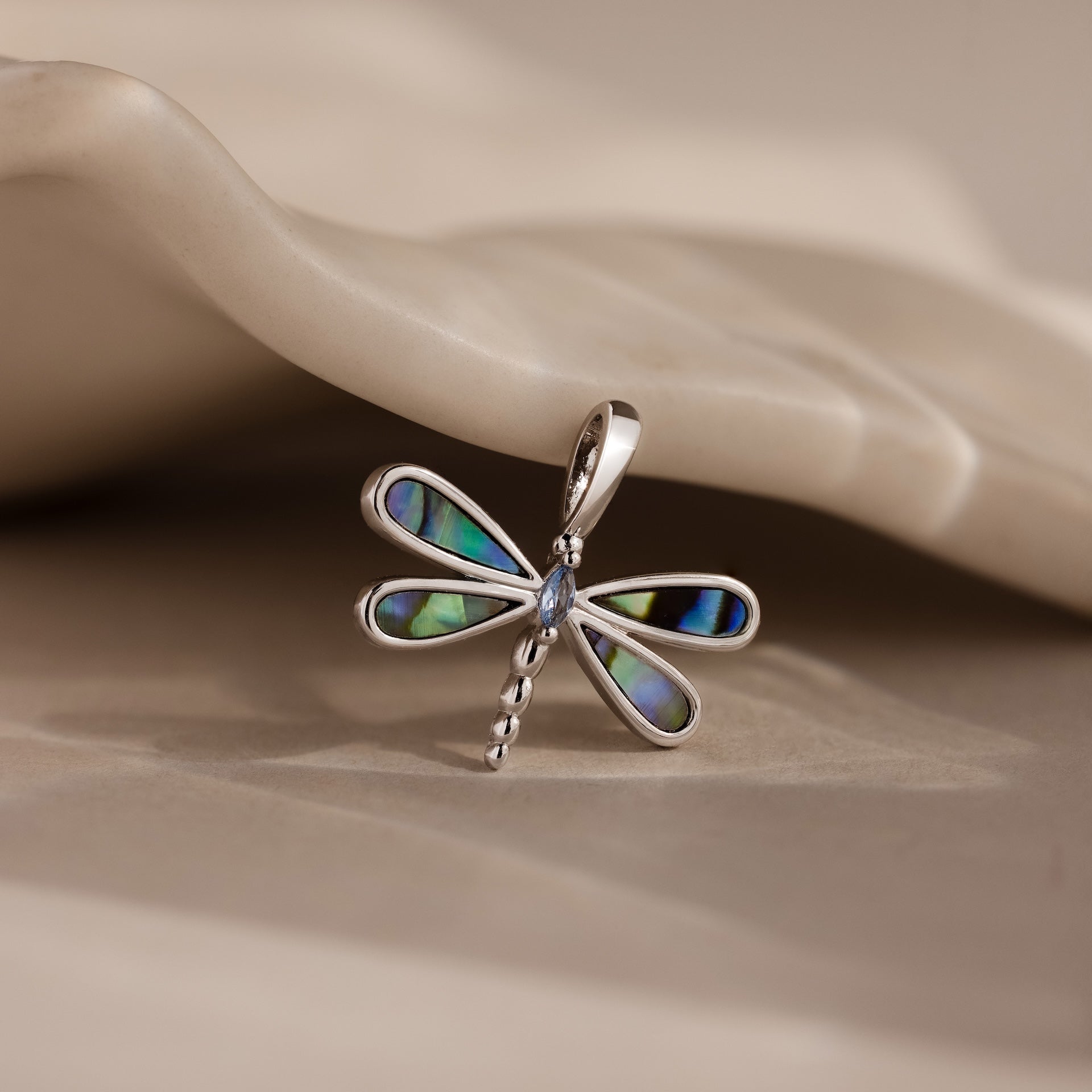 The Abalone Dragonfly Charm in Silver features iridescent wings and rests gracefully on a neutral, wavy surface.