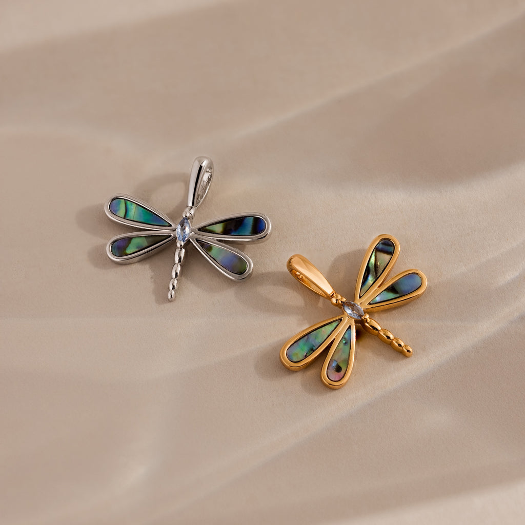 The Abalone Dragonfly Charm in Silver, with iridescent wings, is displayed alongside a gold dragonfly pendant on a beige surface.