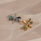 The Abalone Dragonfly Charm in Silver, with iridescent wings, is displayed alongside a gold dragonfly pendant on a beige surface.