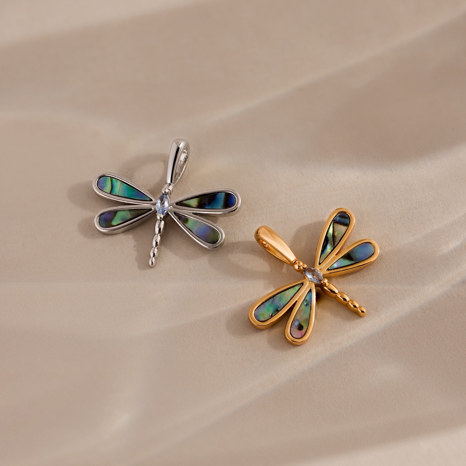 The Abalone Dragonfly Charm in Silver, with iridescent wings, is displayed alongside a gold dragonfly pendant on a beige surface.