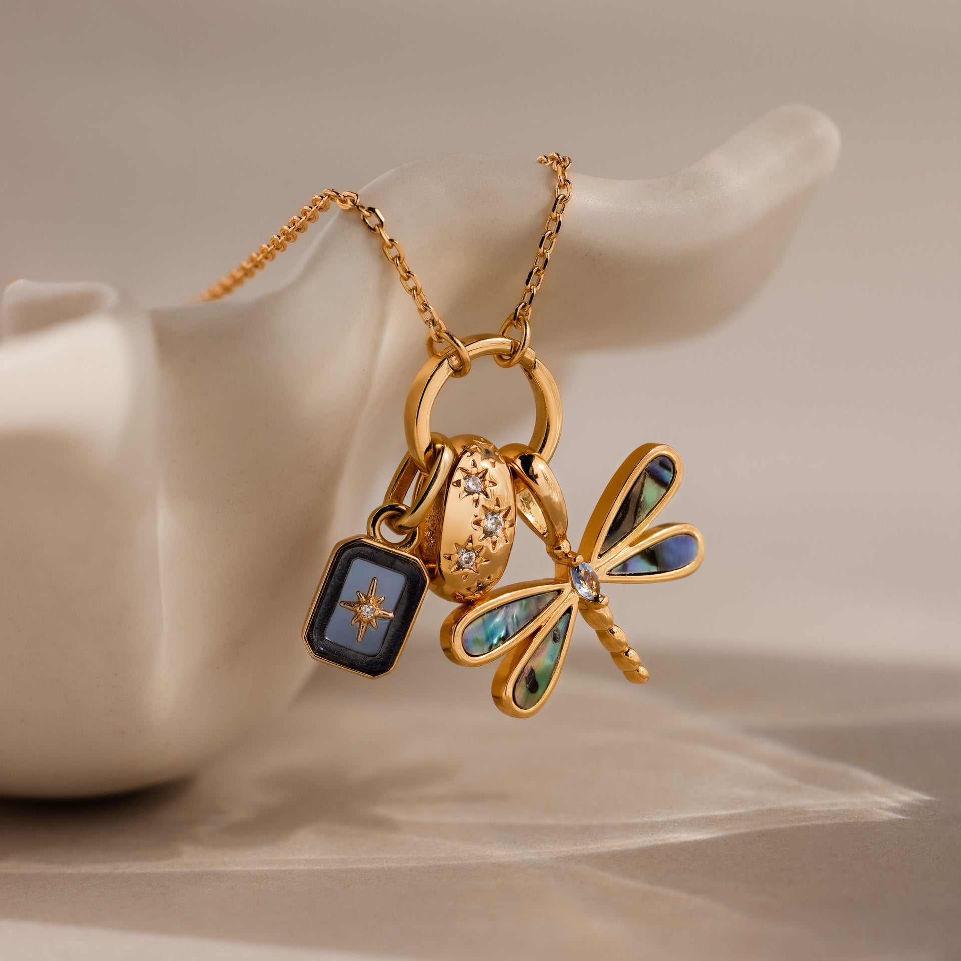 The Abalone Dragonfly Charm in 18K Gold features iridescent wings, a blue accent, and a jeweled ring, displayed on a white ceramic hand.