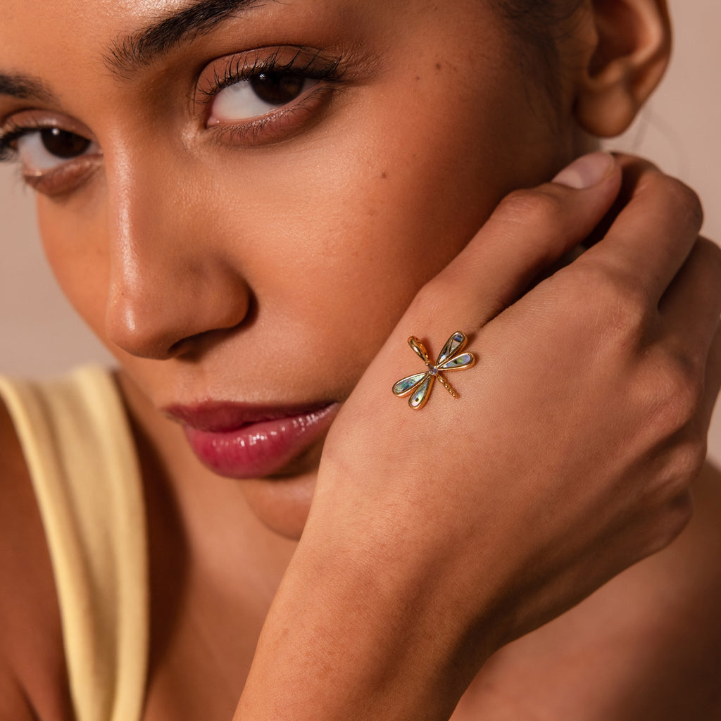 A woman with neutral makeup wears the Abalone Dragonfly Charm in 18K Gold on her hand, showcasing a nature-inspired elegance.