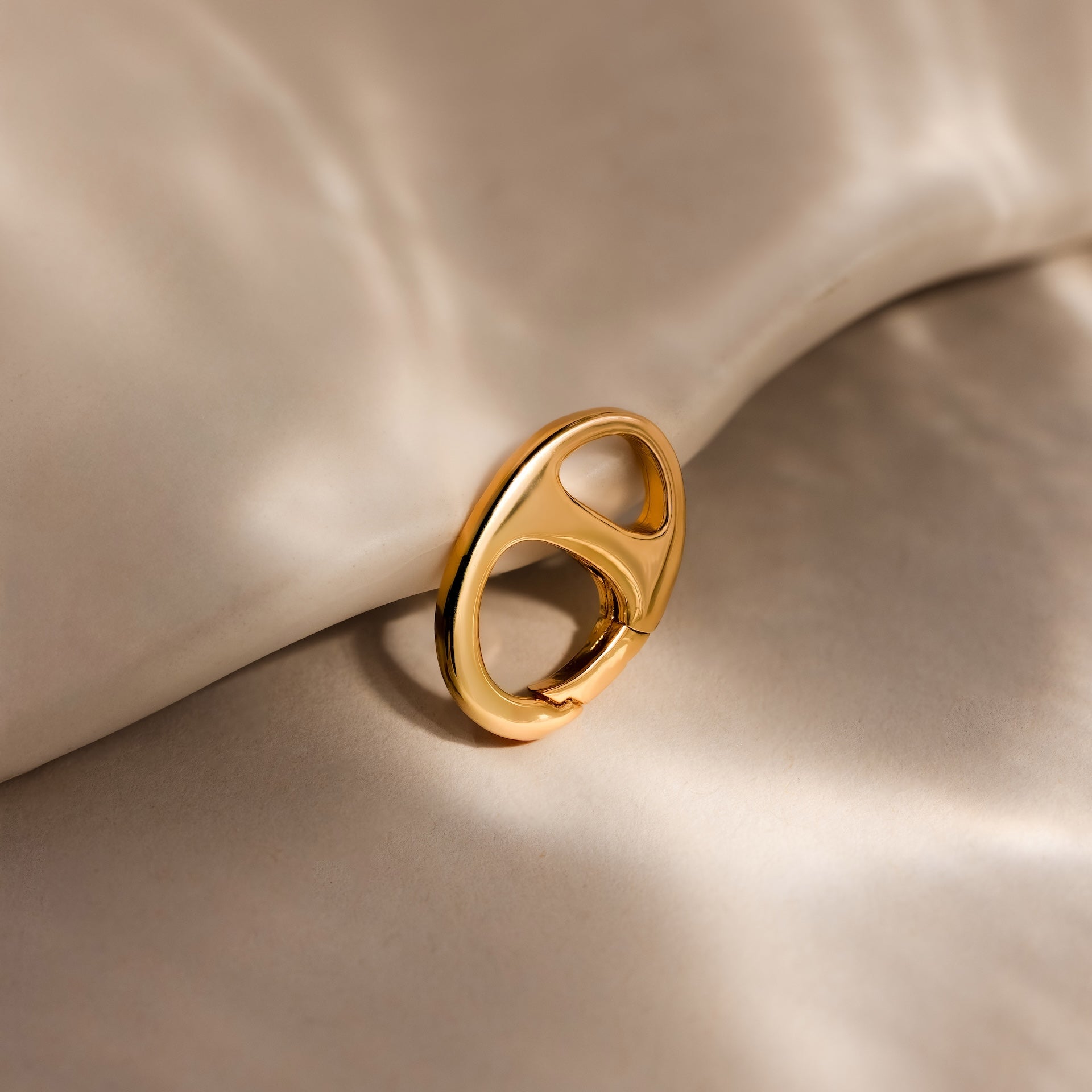 A Connector Clasp in gold with a modern design rests on soft, beige fabric under gentle, diffused lighting.