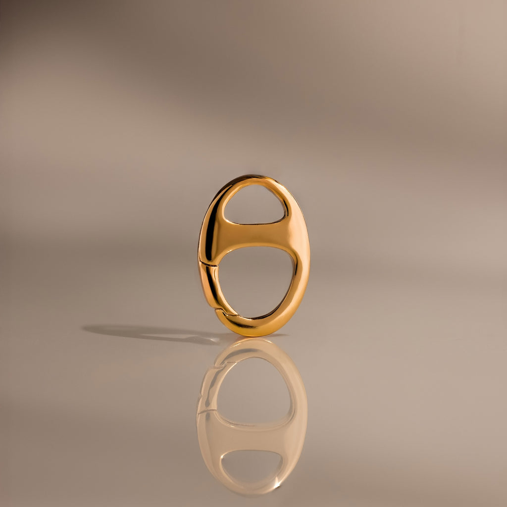 The Connector Clasp features a shiny gold oval design with two cutout sections, standing upright on a reflective surface for a modern, elegant look—ideal for adding a touch of luxury to your décor.