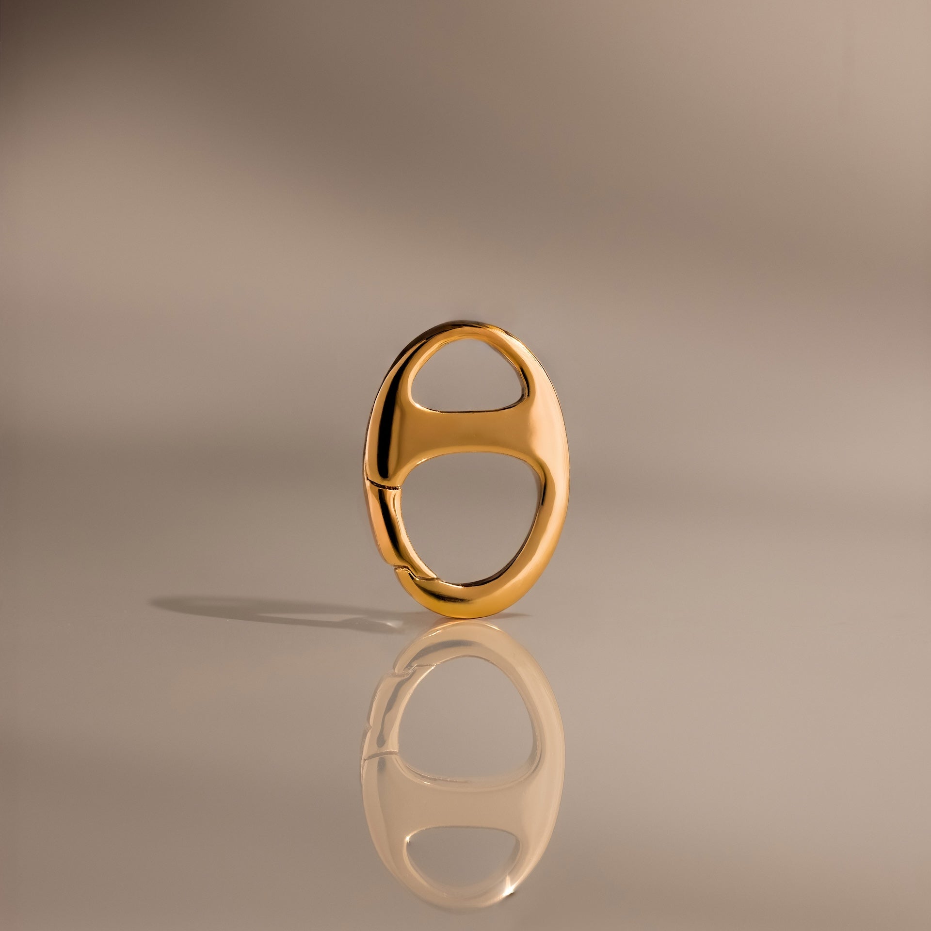 The Connector Clasp features a shiny gold oval design with two cutout sections, standing upright on a reflective surface for a modern, elegant look—ideal for adding a touch of luxury to your décor.