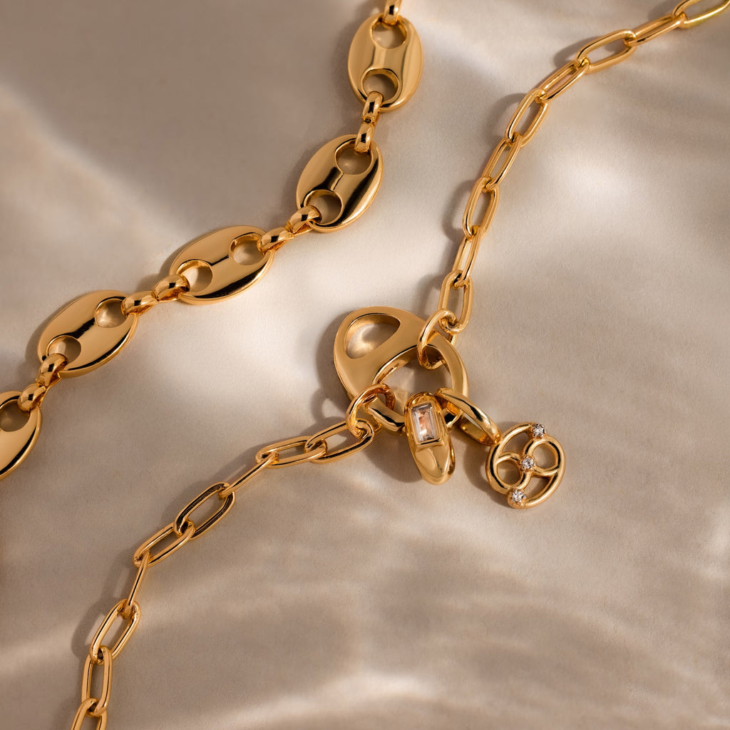 The Connector Clasp gold chain necklaces, featuring unique links and displayed on soft, light fabric, are statement pieces that elevate any outfit with timeless elegance.