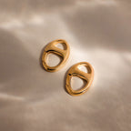 The Connector Clasp: two gold oval hoop earrings with cut-out details rest on a soft, neutral background with gentle shadows.